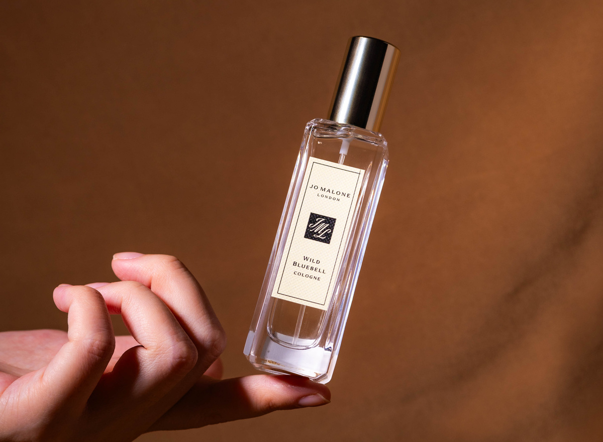 Jo Malone Cologne resting on a female finger, slightly tilted. With a brown silky background. 
